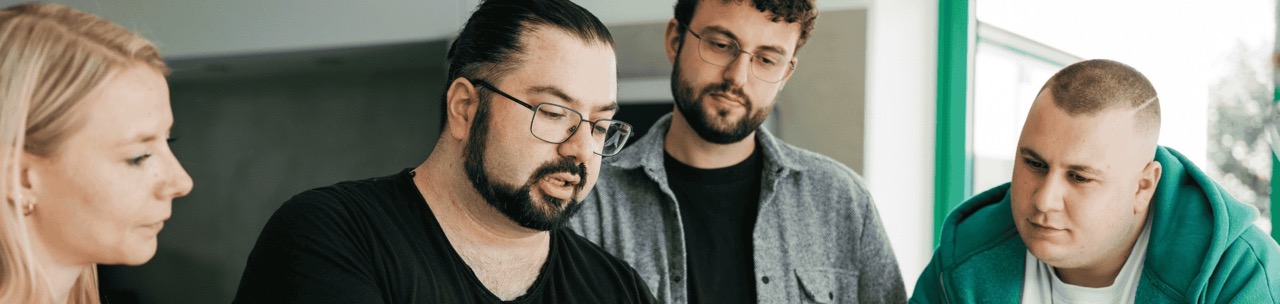 Vier Personen stehen gemeinsam um einen Tisch und schauen auf einen Laptop. In der Mitte erklärt ein Mann mit Brille und schwarzem T-Shirt etwas. Die anderen hören aufmerksam zu.