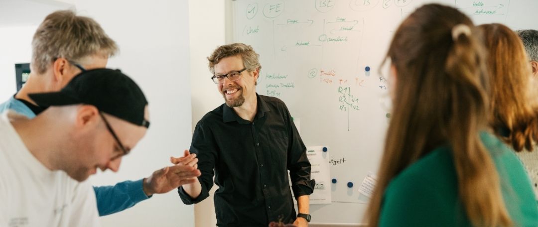 Mehrere Personen vor einem Whiteboard im Gespräch, ein Mann in schwarzem Hemd lacht freundlich.