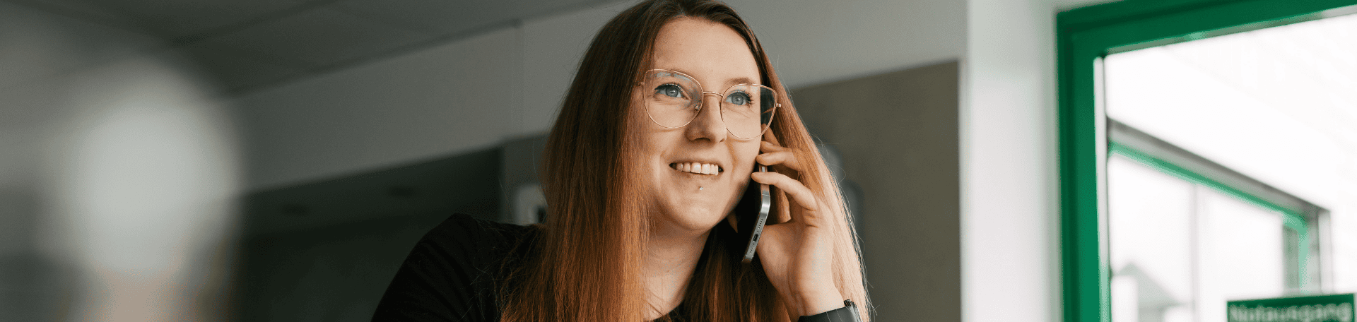Eine Frau mit langen rötlich-blonden Haaren telefoniert lächelnd mit dem Smartphone, vor ihr steht eine weiße Tasse mit Löffel auf dem Tisch.