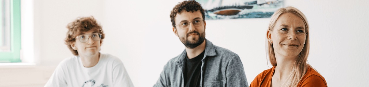 Zwei Männer und eine Frau sitzen an einem Tisch mit Getränken und Laptop und blicken aufmerksam in eine Richtung.