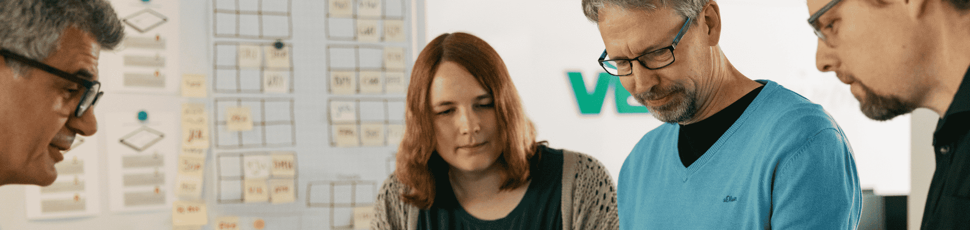 Vier Personen stehen vor einem Whiteboard mit vielen Notizzetteln und tauschen sich aus.